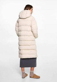 Lunga giacca puffer beige pallido con cappuccio, caratterizzata da un design trapuntato e polsini elasticizzati. Indossata sopra una gonna scura e calzature marroni.