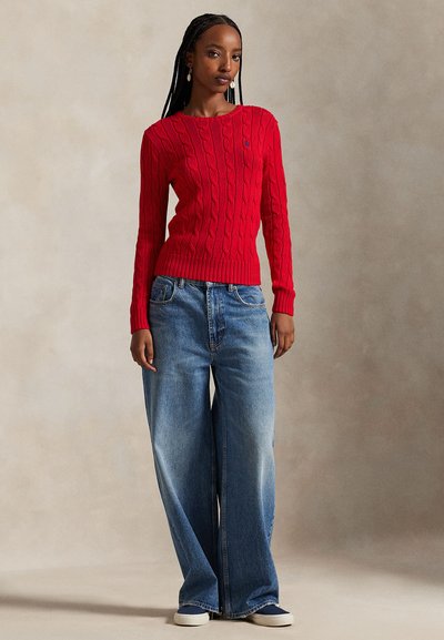 Κόκκινο πουλόβερ με πλέξη καλώδιο και στρογγυλή λαιμόκοψη, συνδυασμένο με φαρδιά μπλε τζιν παντελόνια. Το πουλόβερ διαθέτει ριμπ μανσέτες και τελείωμα.