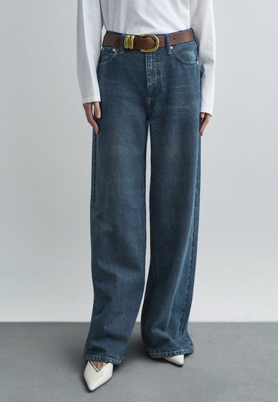 Jean bleu à jambes larges avec texture délavée, doté d'une ceinture marron à boucle dorée. Porté avec des talons blancs à bouts pointus.