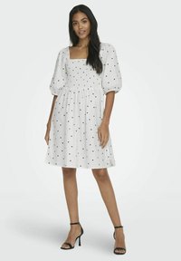 Valge lühike kleit mustade südame mustritega, smockitud bodice, ruudukujuline kaelus ja puhvärv. Ühendatud mustade kõrgete kontsadega sandaalidega.