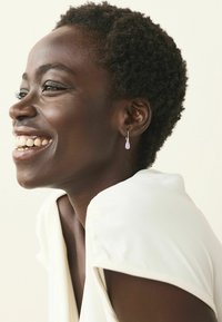 Korvakorut esittävät laventelinväristä pisaramuotoista designia, joka roikkuu hopeaselta koukulta. Aiheella on lyhyt, teksturoitu hiustyyli ja vaalea yläosa.