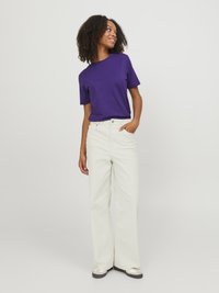 Purple short-sleeve t-shirt, relaxed fit, paired with cream wide-leg jeans. Model has curly hair and pockets in jeans. White sneakers.