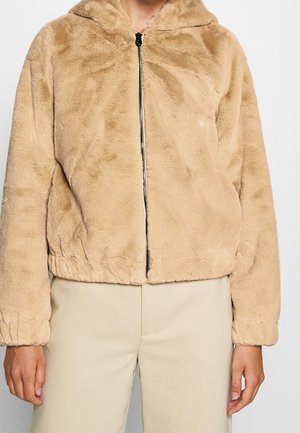 Personne portant une veste marron clair duveteuse à fermeture éclair avec des poignets élastiques, associée à un pantalon beige sur un fond uni.