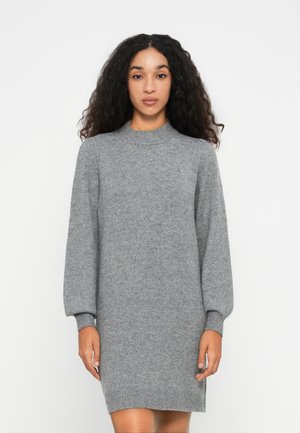 Donna in piedi di fronte, che indossa un vestito maglione grigio a maniche lunghe con polsini e orlo a costine contro uno sfondo bianco semplice.