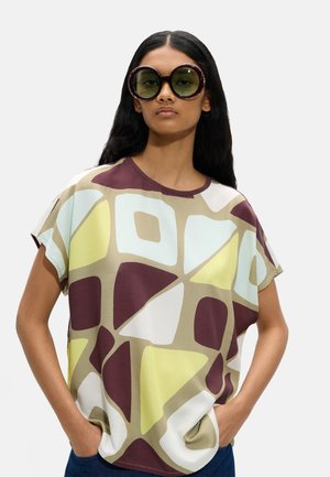 Kobieta w dużych okrągłych okularach przeciwsłonecznych w oprawkach w kolorze żółwia oraz luźnej bluzce z abstrakcyjnymi żółtymi, białymi, beżowymi i bordowymi geometrycznymi kształtami.