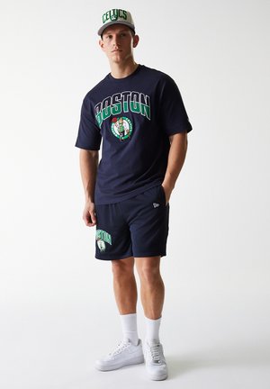 Navy t-shirt with "BOSTON" graphic and Celtic logo, paired with matching navy shorts. White sneakers and a two-tone cap complete the outfit.
