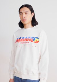 Sweatshirt blanc avec un imprimé graphique coloré et des accents bleus, rouges et jaunes. Poignets et col côtelés, texture douce, coupe classique.