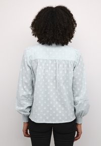 Hellblaue Bluse mit strukturierten Polka-Dots, Puffärmeln und Spitze an den Manschetten. Falten auf der Rückseite verbessern den Fall und das Design.