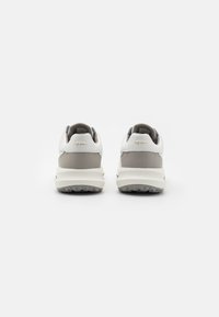 Chaussures de golf blanches avec des accents gris, en matériau synthétique lisse, avec un col rembourré et une semelle en caoutchouc texturé. Logo doré à l'arrière.