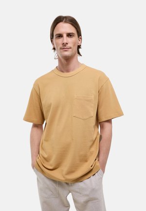 Homme aux cheveux châtains mi-longs, portant une chemise jaune moutarde à manches courtes avec une poche poitrine et un pantalon beige clair, mains dans les poches.