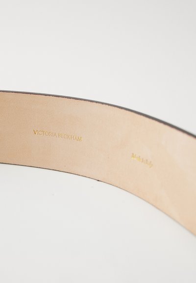 Cinturón de cuero interior con "Victoria Beckham" y "Hecho en Italia" en relieve en dorado, con una superficie lisa en tono marrón claro y borde oscuro.