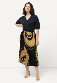 Marinblå V-ringad topp i kombination med en plisserad kjol i guld och svart, med ett djärvt abstrakt mönster. Modellen har på sig beige klackar.