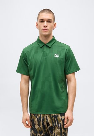 Modèle masculin aux cheveux courts portant un polo vert à col avec un petit logo blanc et un pantalon à motif camouflage sur un fond uni.