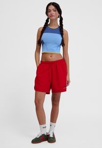 Blå kort topp med gula detaljer och geometriska mönster, kombinerad med röda shorts med dragsko. Modellen bär gröna och röda sneakers med vita strumpor.