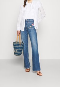 Jean bleu brodé de fleurs avec une jambe évasée, associé à une chemise blanche, un t-shirt blanc uni en dessous, et un sac tissé bleu.