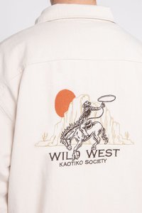Beige stoffen shirt met een geborduurd ontwerp van een cowboy op een paard, met een rode zon en de tekst "WILD WEST KAOTIKO SOCIETY" eronder.