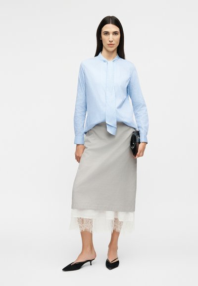 Γυναίκα στέκεται φορώντας μπλούζα με μακριά μανίκια σε ανοιχτό γαλάζιο με ρίγες, γκρι midi φούστα με λευκή δαντέλα στο τελείωμα, μαύρα παπούτσια με χαμηλό τακούνι και κρατώντας μαύρο τσαντάκι χειρός.