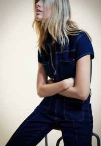 Mörk denimbyxdress med korta ärmar, blixtlåsförclosure, två bröstfickor och detaljerad sömnad. Modellen sitter med armarna i kors.