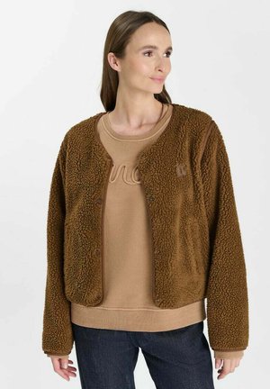 Donna che indossa una giacca in pile marrone con trama sopra una felpa beige e jeans scuri, guarda leggermente verso destra su uno sfondo bianco.
