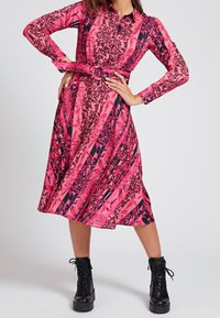 Abito a maniche lunghe in un audace rosa con motivi floreali, cinturato in vita e con colletto. Indossato con stivaletti alla caviglia neri con lacci.