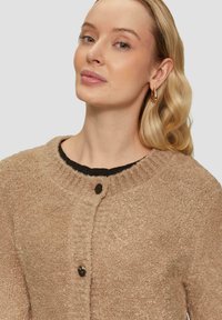 Beżowy sweter z teksturowanej dzianiny, z prążkowanym kołnierzem, okrągłym dekoltem i pojedynczym czarnym zapięciem z przodu.