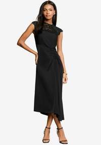 Vestito nero con maniche a cappuccio in pizzo, dettaglio arricciato in vita, orlo asimmetrico e tessuto liscio. Indossato con tacchi neri a strappo.