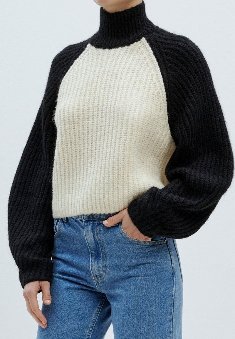 Chunky gebreide trui met een hoge kraag, zwart en crème kleuren, geribbelde textuur en een ontwerp met verlaagde schouders. Gepaard met blauwe jeans.