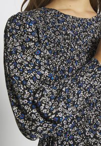 Musta kukkakuvioinen mekko, jossa on smokkaukset, pienet valkoiset, siniset ja punaiset kukat kankaalla ja kerätyt pitkät hihat.