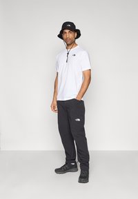 Άσπρο κοντομάνικο πουκάμισο με μαύρο λογότυπο, μαύρο καπέλο τύπου κάπα, μαύρο παντελόνι και μαύρα παπούτσια. Απλός σχεδιασμός με άνετη εφαρμογή.