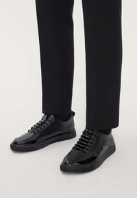 Zapatillas negras con un upper de cuero liso y acentos de charol, punta redonda y suela de goma plana, combinadas con pantalones negros ajustados.