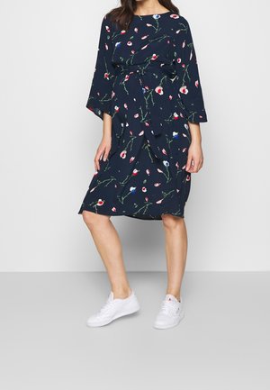 Kvinde iført marineblå knælængde kjole med blomsterprint og 3/4 ærmer samt hvide sneakers, stående mod en ensartet lys baggrund.