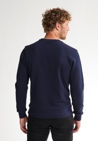 Felpa blu navy con scollo a girocollo, tessuto testurizzato e polsini a costina. Mostrata da dietro con silhouette aderente e dettagli minimi.