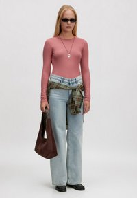 Rosa gerippter Langarmshirt mit hellblauen weit geschnittenen Jeans, grün-braun kariertes Hemd, das um die Taille gebunden ist, braune Wildleder-Handtasche, schwarze Schuhe.
