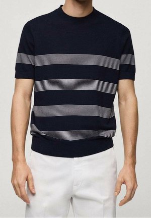 Homme portant un polo en maille bleu marine à manches courtes avec des rayures horizontales blanches, associé à un pantalon blanc.