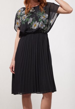Vestito nero plissettato con sovrapposizione trasparente floreale, caratterizzato da margherite bianche e accenti verdi. Vita elastica e maniche corte e larghe.