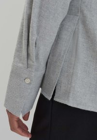 Haut gris à manches longues en tissu doux, présentant une fente sur le côté et un détail de bouton aux poignets. L'ourlet est légèrement arrondi avec des bords bruts.