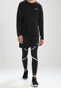 Schwarzes langärmeliges Nike-Shirt mit Logo, kombiniert mit schwarzen Shorts und formschlüssigen schwarzen Leggings mit weißen Akzenten sowie schwarzen Sportschuhen.