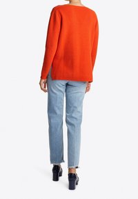 Pull orange en maille à coupe décontractée, manches longues et fentes latérales, associé à un jean bleu clair à jambes droites et des talons épais noirs.