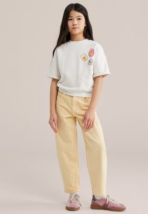 Fată tânără cu păr lung și închis la culoare, purtând un tricou alb cu mâneci scurte și patch-uri colorate, pantaloni bej și adidași roz, stând pe un fundal neutru.