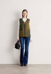 Πράσινο καπιτονέ γιλέκο με στρογγυλή αγκράφα, τσέπες μπροστά, φορεμένο πάνω από λευκό πουκάμισο με μακριά μανίκια και συνδυασμένο με μπλε τζιν παντελόνι με καμπάνα.