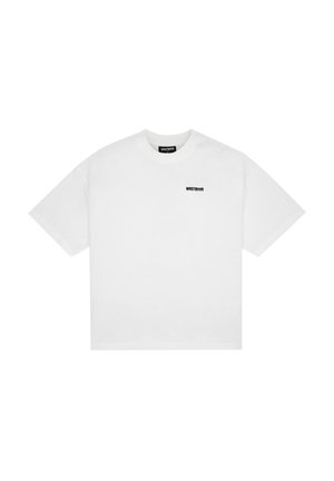 Joven con camiseta blanca holgada con pequeño texto negro en el pecho y pantalones de color lila claro, mirando de lado sobre fondo liso.