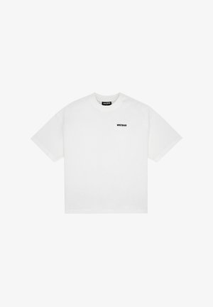 Młody mężczyzna w luźnej białej koszulce z małym czarnym napisem na piersi i jasnofioletowych spodniach, patrzący w bok na jednolitym tle.