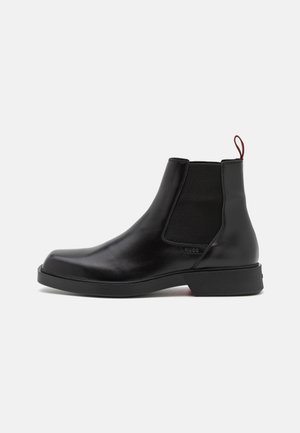 Bottes Chelsea en cuir noir avec panneaux latéraux élastiques, bout rond et semelle en caoutchouc texturée. Elles comportent une languette de tirage rouge à l'arrière.