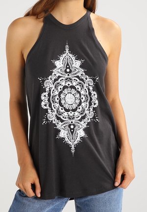 Frau trägt ein ärmelloses schwarzes Neckholder-Top mit einem filigranen weißen, symmetrischen Mandala-Design auf der Vorderseite.