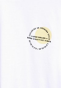 Bílé bavlněné tričko s kruhovou grafikou a textem: "Prozkoumávání neznámého oceánu," "Zůstaňte v klidu" a "Hledejte pozitivní vibrace."