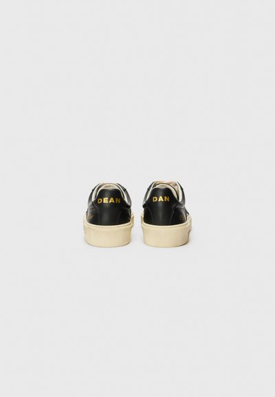 Zapatillas negras de cuero con suelas blancas, con "DEAN" y "DAN" impresos en amarillo en los talones y un collarín acolchado para mayor comodidad.