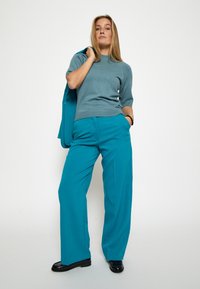 Tamsiai mėlynas megztinis su trumpomis rankovėmis, derinamas su plačiomis tamsiai mėlynomis kelnėmis. Modelis laiko tamsiai mėlyną švarką, o ant kojų turi juodas lakinės odos batelius.