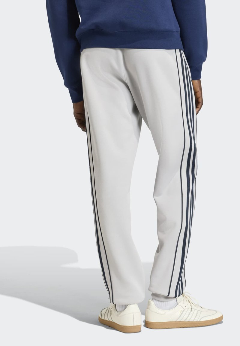 Personne portant un pantalon de survêtement blanc avec des rayures verticales foncées sur les côtés, des chaussettes blanches, des baskets blanches avec semelles en gomme et un sweat-shirt bleu marine.
