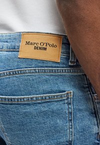 Svetlomodré džínsové nohavice s hnedou koženou značkou s nápisom "Marc O'Polo DENIM." Obsahujú detaily šitia a zadné vrecká.
