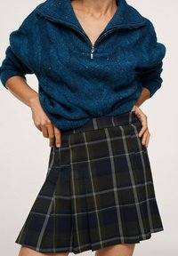 Teal kabelgebreide trui met hoge kraag en ritssluiting aan de voorkant, gecombineerd met een geplooide zwart-groene ruitjesrok met grijze accenten.